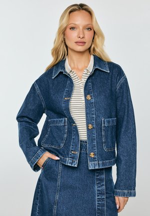 Blondynka ma na sobie niebieską dżinsową kurtkę z przednimi kieszeniami i guzikami, pod którą znajduje się pasiasta koszula z kołnierzykiem, oraz dopasowaną dżinsową spódnicę.