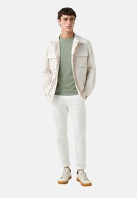 Lichtbeige jas met meerdere voorzakken, groene trui, witte jeans en beige sneakers met rubberen zolen, staande tegen een witte achtergrond.