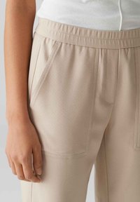 Beige taps toelopers met een elastische tailleband, voorzien van diagonale zakken en een gestructureerde stof, gericht op duurzaamheid en comfort.