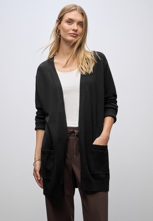 Cardigan noir à coupe ample, avec un devant ouvert et des poignets côtelés. Il comporte deux poches avant et une texture douce. Porté sur un haut blanc.