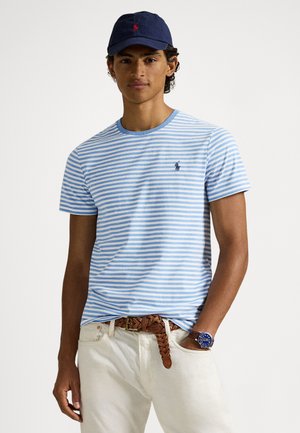 Μπλε και λευκή ριγέ μπλούζα με στρογγυλή λαιμόκοψη, κοντά μανίκια και ένα μικρό λογότυπο, συνδυασμένη με ανοιχτόχρωμο παντελόνι και καφέ πλεκτή ζώνη.