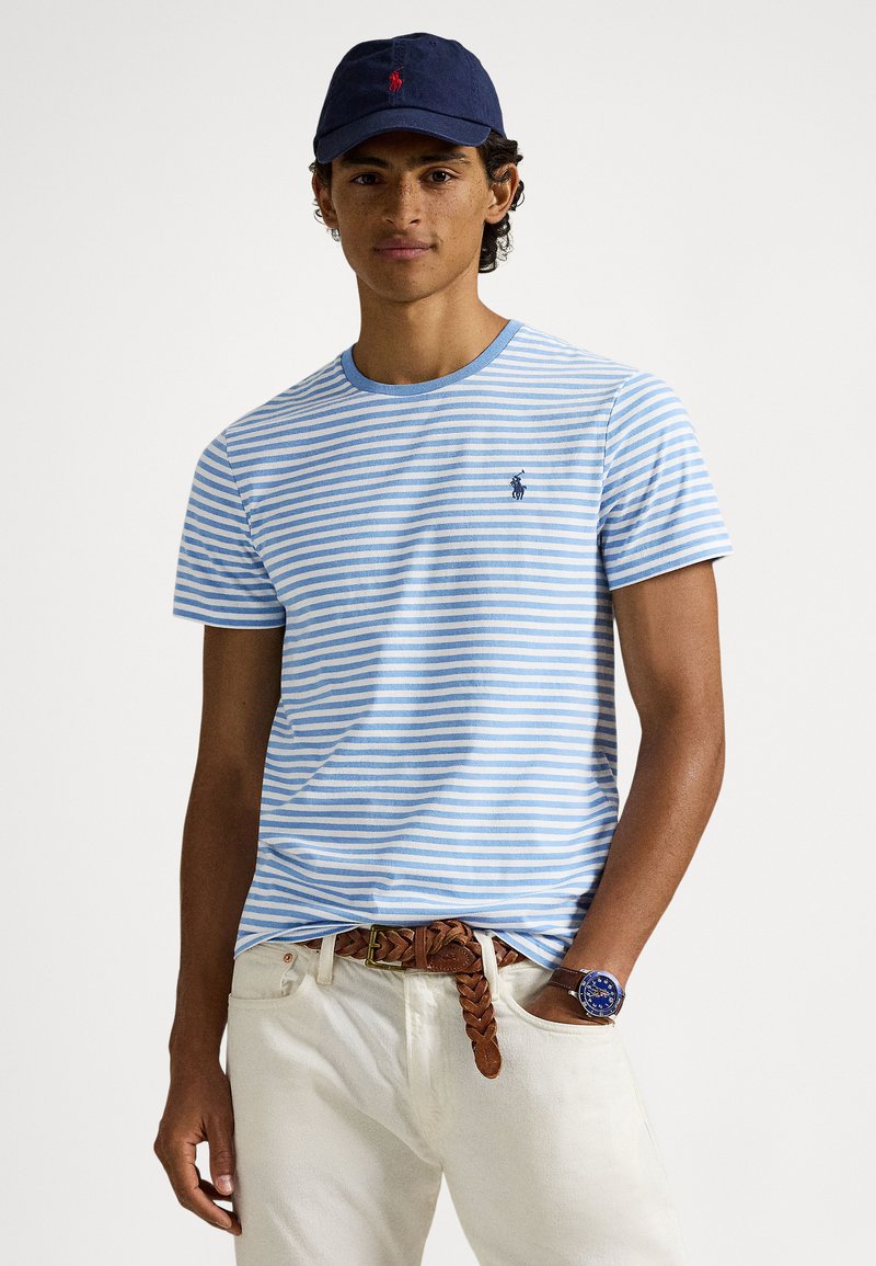 Μπλε και λευκή ριγέ μπλούζα με στρογγυλή λαιμόκο�ψη, κοντά μανίκια και ένα μικρό λογότυπο, συνδυασμένη με ανοιχτόχρωμο παντελόνι και καφέ πλεκτή ζώνη.