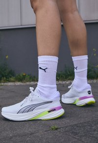 Zapatillas de running blancas con un diseño elegante, acentos negros, detalle en la suela en púrpura y verde, combinadas con calcetines blancos de canalé que presentan un logo negro.