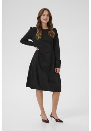 Robe noire à manches longues avec une coupe décontractée, présentant des détails froncés à la taille et un décolleté rond. Associée à des chaussures à talons hauts bordeaux.
