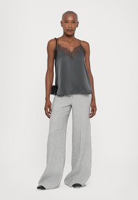 Woman wearing a black lace-trimmed camisole, wide gray pinstriped pants, black shoes, and a black shoulder bag, standing on a white background.