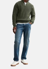 Pullover lavorato a maglia verde oliva con colletto a mezza cerniera, abbinato a jeans azzurri e sneakers bianche con dettagli beige.