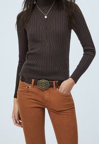 Dunkelbrauner, gerippter Rollkragenpullover kombiniert mit rostfarbenen Cordhosen, ergänzt durch einen schwarzen Gürtel mit einer floral geprägten Metallschnalle.