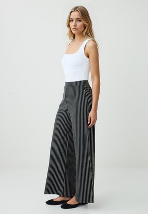 Una modella posa indossando un top bianco aderente e pantaloni a gamba larga a righe bianche e nere, sullo sfondo di un ambiente neutro.