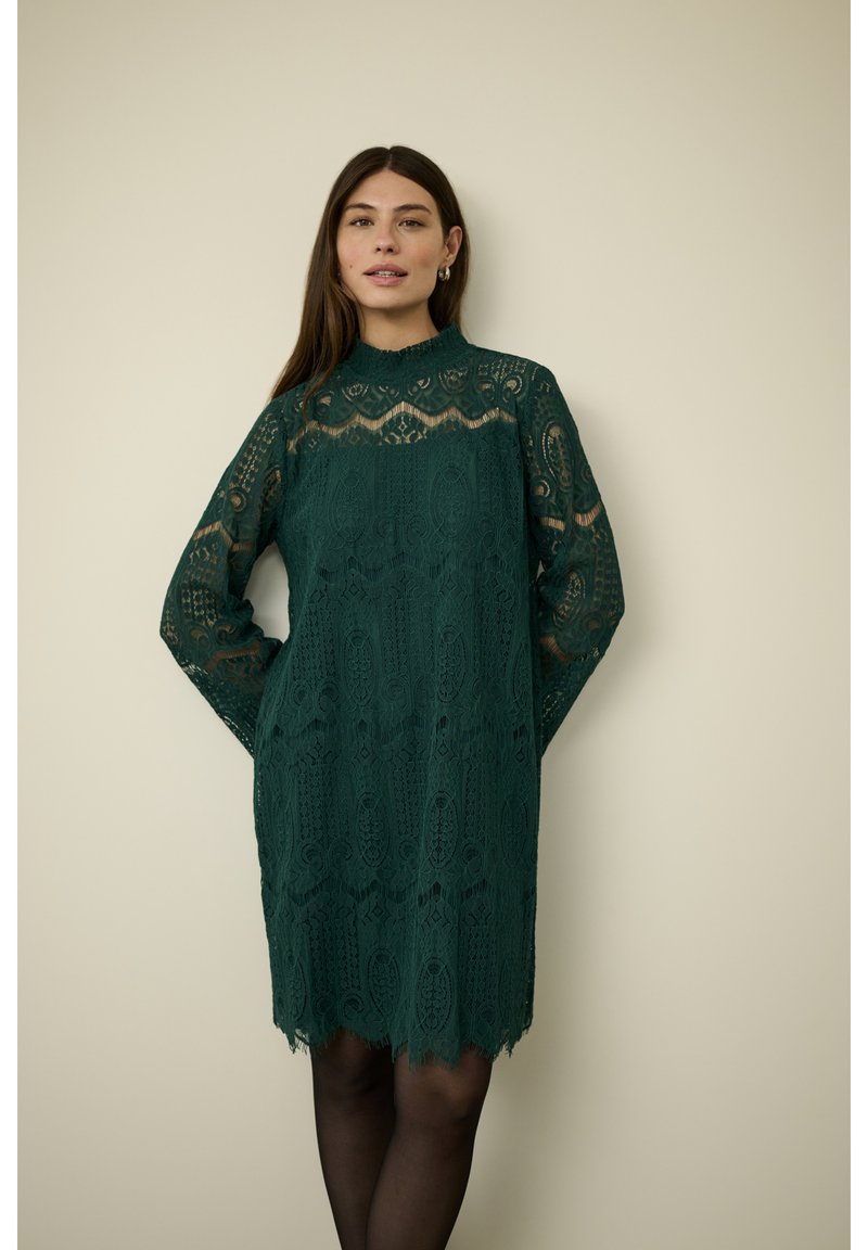 Tmavě zelené krajkové šaty s dlouhými rukávy, vysokým výstřihem, složitými vzory a zvlněným lemem. Nosené přes černé punčocháče.