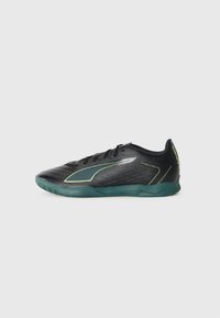 Zapatilla de fútbol sala negra y verde oscuro con cordones, parte superior texturizada y texto "ULTRA" en el talón, mostrada de lado sobre un fondo blanco.