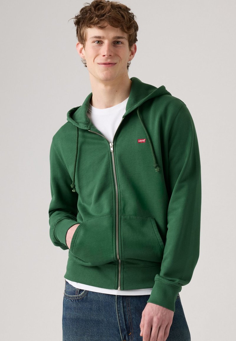 Felpa verde con zip realizzata in tessuto morbido, dotata di una tasca frontale, cappuccio con cordino e piccolo logo rosso sul lato sinistro del petto.