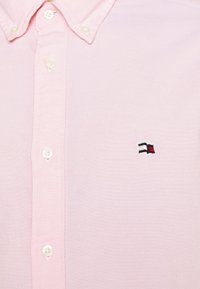 Camicia rosa chiaro con bottoni bianchi e colletto, presenta un piccolo logo ricamato di una bandiera in nero, bianco e rosso sul petto.
