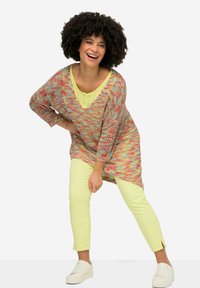 Multikleurige gebreide trui met een V-hals en driekwartmouwen, gecombineerd met lichtgele slim-fit broek en witte sneakers.