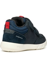 Baskets imperméables bleu marine avec dessus en daim, bande velcro, languette de talon rouge contrastante et semelle en caoutchouc blanc avec un detailing texturé.
