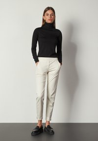 Schwarzer Rollkragenpullover, figurbetontes Design; cremefarbene, taillierte Hose mit Seitentaschen, schmal zulaufendes Bein; schwarze Slipper mit dicker Sohle.