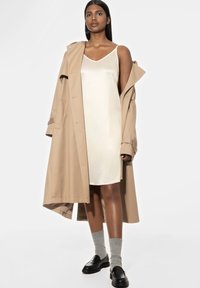 Beige trenchcoat over een crèmekleurige jurk. Grijze sokken en zwarte loafers maken de look af. Glad textuur en minimalistisch ontwerp.