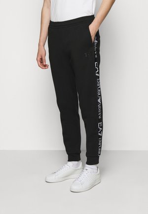 Pantalon de jogging noir en mélange de coton, avec une coupe fuselée, une taille élastique et un imprimé logo blanc sur le côté. Associé à des baskets blanches.