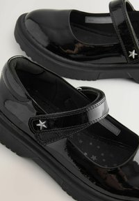 Pantofi Mary Jane din piele lăcuită neagră, cu curea Velcro, având un accent în formă de stea argintie și o talpă interioară căptușită cu modele de stele.