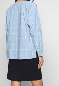 Chemise bleu clair à manches longues avec un motif à carreaux, portée sur une jupe noire. La chemise est de coupe décontractée avec des manches retroussées.