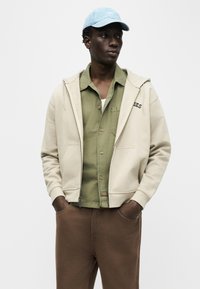 Muž nosiaci svetlomodrú čiapku, béžovú mikinu na zips, olivovo zelenú košeľu na gombíky a hnedé nohavice, stojaci s rukami v vreckách.