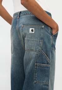 Μπλε τζιν παντελόνι cargo με ψηλή μέση, πολλαπλές τσέπες και λευκή ετικέτα με το σήμα στο πίσω μέρος, με ξεβαμμένη υφή.