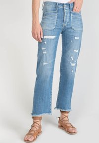 Jeans déchirés bleu clair à coupe droite, avec des ourlets effilochés et plusieurs détails vieillissants. Associés à des sandales à lanières beiges.