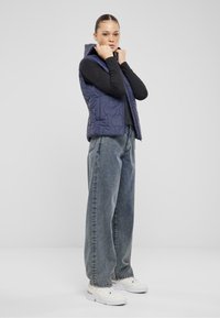 Marinblå quiltad väst med huva, bärs över en svart långärmad topp, tillsammans med ljusblå, löst sittande jeans och vita sneakers.