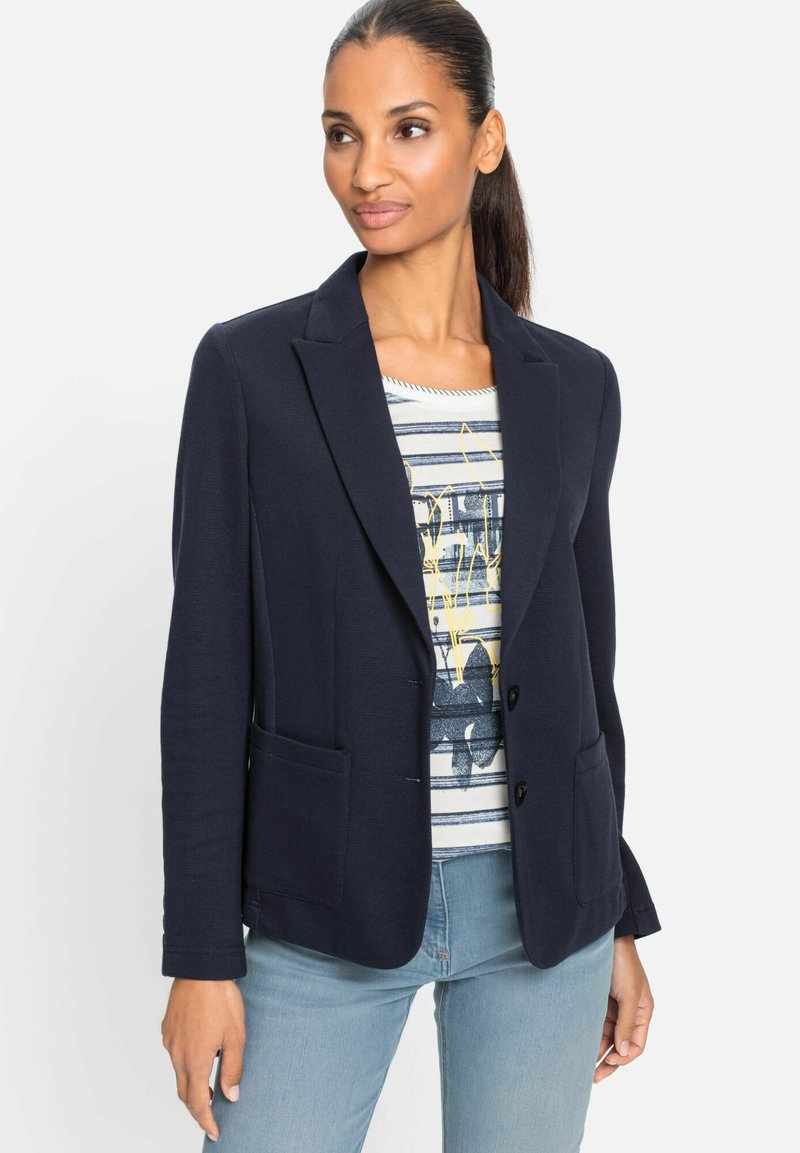 Navy blazer with a fitted design, single-button closure, two front pockets, and a smooth texture, worn over a printed top and jeans.