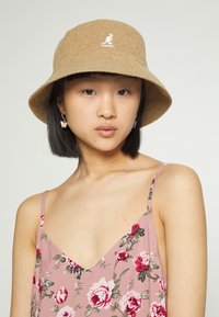 Chapéu de balde castanho feito de tecido texturizado com um logótipo bordado. Modelo veste um vestido floral rosa com alças finas.