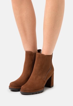 Hnědé semišové kotníkové boty na blokovém podpatku, s bočními elastickými panely a zaoblenou špičkou. Texturovaný povrch a odolná podrážka.