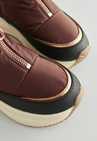 Sneaker in tessuto marrone con chiusura a zip, dettagli dorati, pannelli laterali neri testurizzati e una suola spessa di colore crema.