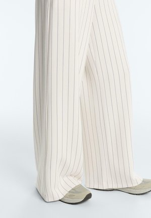 Wijd uitlopende crèmekleurige broek met dunne verticale zwarte strepen, gedragen met beige en grijze sneakers op een lichte achtergrond.