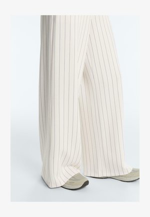 Pantaloni crem cu croială largă și dungi verticale subțiri negre, purtați cu adidași bej și gri pe un fundal deschis.