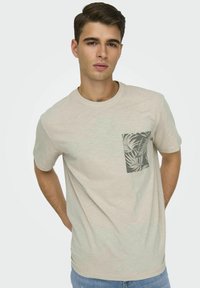 T-shirt di cotone color beige chiaro con taschino grigio a motivo foglie tropicali, scollo rotondo e maniche corte. Vestibilità casual.