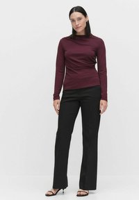 Bordeaux lange mouwen top met een gedrapeerd ontwerp, gecombineerd met zwarte rechte broek en open teen zwarte hakken op een neutrale achtergrond.
