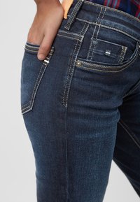 Jeans de mezclilla azul oscuro con un diseño ajustado, con costuras amarillas en contraste, bolsillos frontales y un pequeño detalle de logo en la parte trasera.