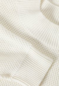 Gros plan sur un pull en tricot couleur crème montrant des motifs de points texturés ainsi qu'un col et des poignets côtelés.