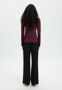 Pikkade lokkis juustega naine seisab seljaga, seljas kohandatud tumepruun pika varrukaga särk ja laiad mustad püksid, mille allääred on kitsendatud.