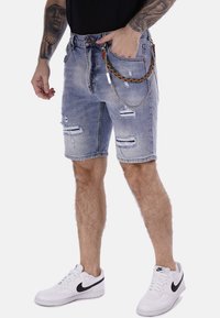 Shorts de mezclilla en azul claro con detalles desgastados, que incluyen bolsillos y una cadena decorativa. Combinados con zapatillas blancas.