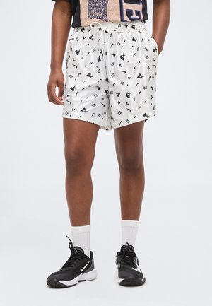 Személy, aki fehér sport rövidnadrágot visel fekete absztrakt mintákkal, fehér bokazoknit, és fekete Nike tornacipőt, zsebre tett kézzel áll.