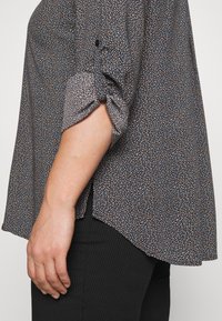 Femme portant un chemisier à motifs avec les manches retroussées et un pantalon noir côtelé, vue de profil montrant son bras et sa main détendus le long du corps.