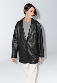 Zwarte leren blazer met een enkele knoopsluiting, gerevers met inkeping en twee voorzakken. Draagt over een grijze top en gecombineerd met lichte broek.