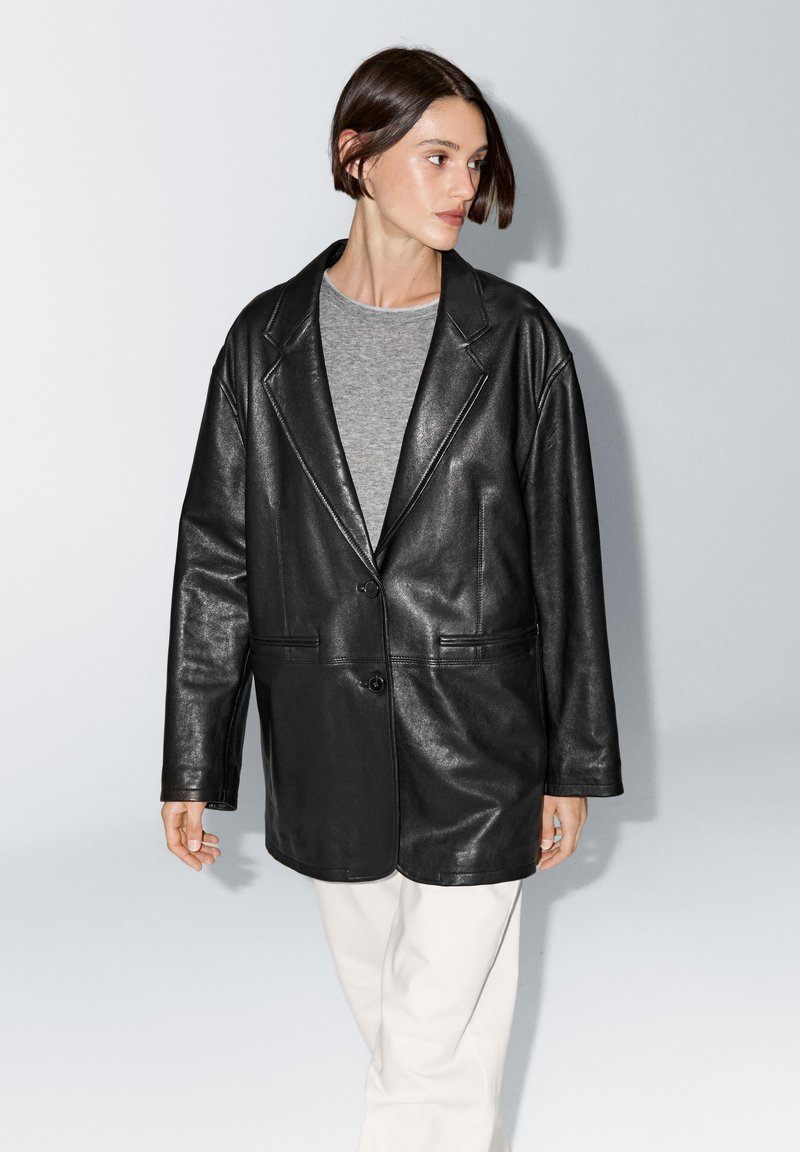 Zwarte leren blazer met een enkele knoopsluiting, gerevers met inkeping en twee voorzakken. Draagt over een grijze top en gecombineerd met lichte broek.