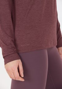 L�ångärmad atletisk tröja i bordeaux, tillverkad av snabbtorkande tyg. Har en platta söm och en figurnära design som matchas med lila leggings.