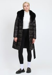 Giovane donna che indossa un cappotto imbottito nero con colletto di pelliccia, dolcevita bianco, leggings neri, calzini bianchi e mocassini neri con dettaglio di catena dorata.