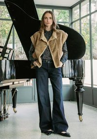 Gilet en peluche brun avec col en fourrure sur un chemisier à motifs navy, associé à un jean foncé évasé et des chaussures noires pointues, se tient à côté d'un piano.