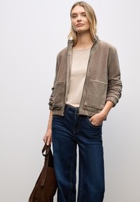 Giacca in corduroy marrone con zip frontale e due tasche anteriori, abbinata a un top beige e jeans a gamba larga blu. Borsa a spalla marrone tenuta in mano.
