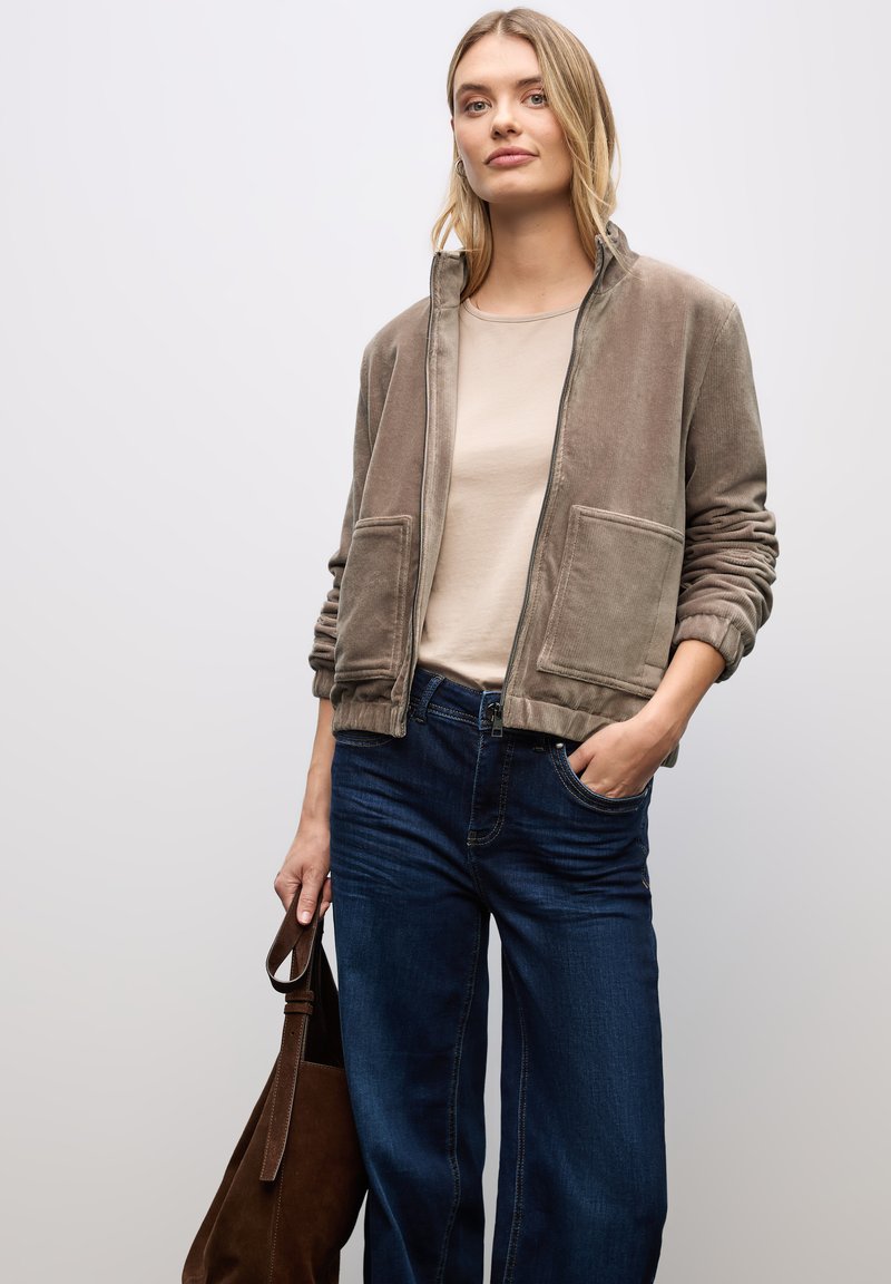 Giacca in corduroy marrone con zip frontale e due tasche anteriori, abbinata a un top beige e jeans a gamba larga blu. Borsa a spalla marrone tenuta in mano.