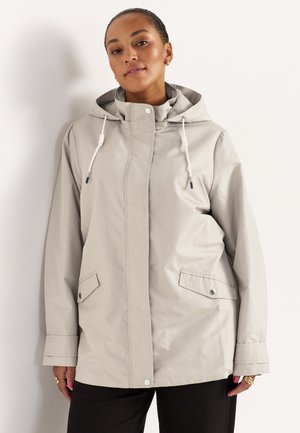 Frau trägt eine hellgraue Regenjacke mit Kapuze, Kordelzügen und Fronttaschen mit Klappe, steht vor weißem Hintergrund.