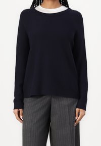 Eine Frau trägt einen dunkelblauen Pullover über einem weißen Hemd, kombiniert mit grauen, gestreiften, weit geschnittenen Hosen, und steht vor einem weißen Hintergrund.